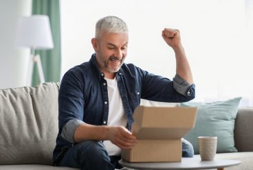 Happy senior man customer checking mail box, raising fists up and screaming, sitting on sofa at home, copy space. Emotional elderly man opening delivery, checking order. Delivery concept